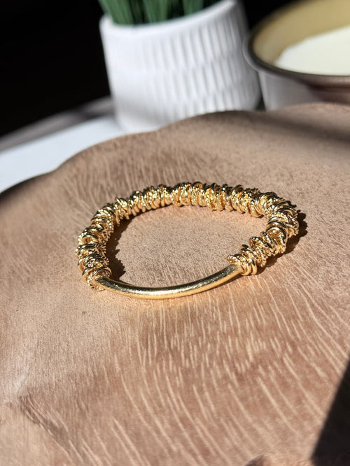 A textured brushed gold bangle bracelet with a polished straight bar, resting on a brown wooden surface with a blurred white vase in the background.