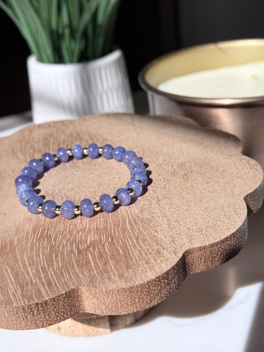A smooth rondelle beaded stretch bracelet made of round lavender-blue stones separated by a gold filled spacer bead, displayed on a scalloped wooden stand.