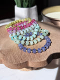 A set of rondelle gemstone beaded bracelets arranged on a scalloped wooden tray. The bracelets feature beads in yellow-green, pink, mint green, lavender, and periwinkle, separated by small gold filled spacer beads.
