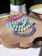 A set of rondelle gemstone beaded bracelets arranged on a scalloped wooden tray. The bracelets feature beads in yellow-green, pink, mint green, lavender, and periwinkle, separated by small gold filled spacer beads.