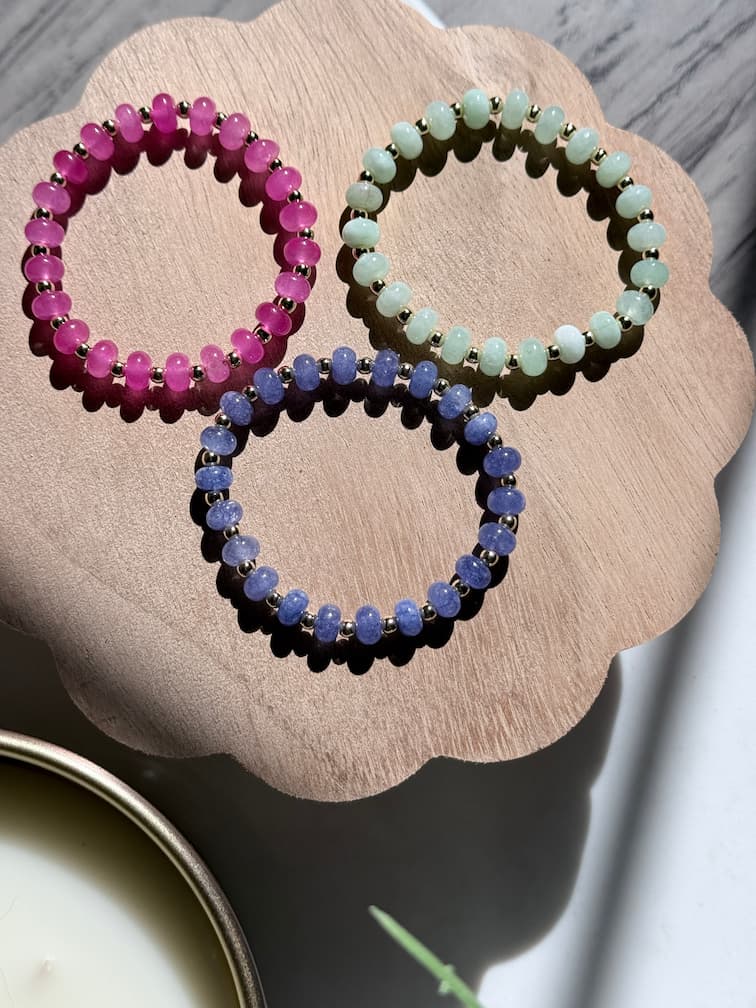 Three beaded stretch bracelets in pink, light green, and blue-purple are arranged on a scalloped wooden tray. The bracelets feature smooth rondelle gemstone beads separated by small gold filled spacers, photographed in bright natural light with soft shadows.