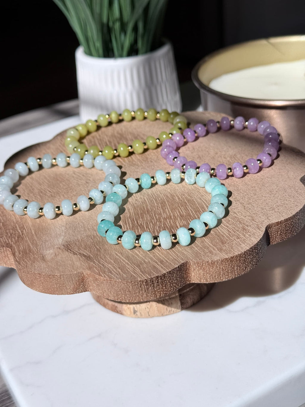 Four beaded stretch bracelets in pastel shades of mint green, lavender, light blue, and olive yellow, displayed on a scalloped wooden stand.  The bracelets feature smooth rondelle gemstone beads separated by small gold filled spacers, photographed in bright natural light with soft shadows. A white ceramic pot and a bowl are softly blurred in the background.