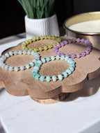 Four beaded stretch bracelets in pastel shades of mint green, lavender, light blue, and olive yellow, displayed on a scalloped wooden stand.  The bracelets feature smooth rondelle gemstone beads separated by small gold filled spacers, photographed in bright natural light with soft shadows. A white ceramic pot and a bowl are softly blurred in the background.