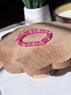 A pink beaded bracelet with small gold twist spacer beads, displayed on a scalloped wooden stand.