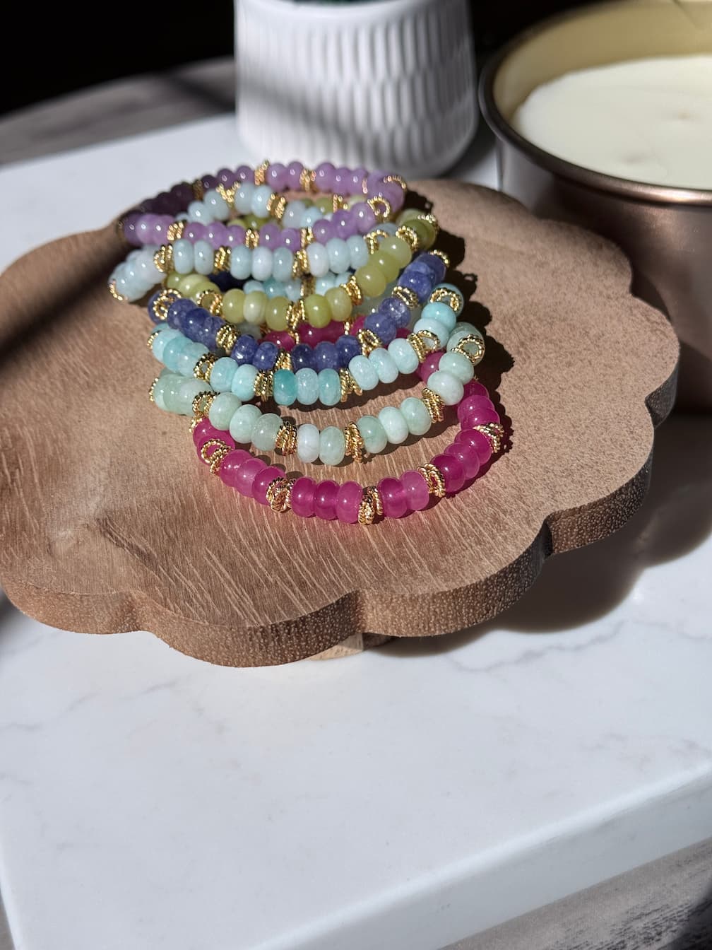 A set of rondelle gemstone beaded bracelets arranged on a scalloped wooden tray. The bracelets feature beads in yellow-green, pink, mint green, lavender, and periwinkle, separated by small gold twist spacer beads.