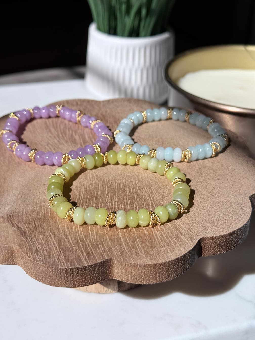 Three beaded stretch bracelets in pastel purple, light blue, and pale yellow-green, each accented with gold twist spacer beads, arranged on a scalloped wooden tray.