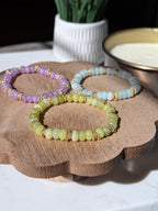 Three beaded stretch bracelets in pastel purple, light blue, and pale yellow-green, each accented with gold twist spacer beads, arranged on a scalloped wooden tray.