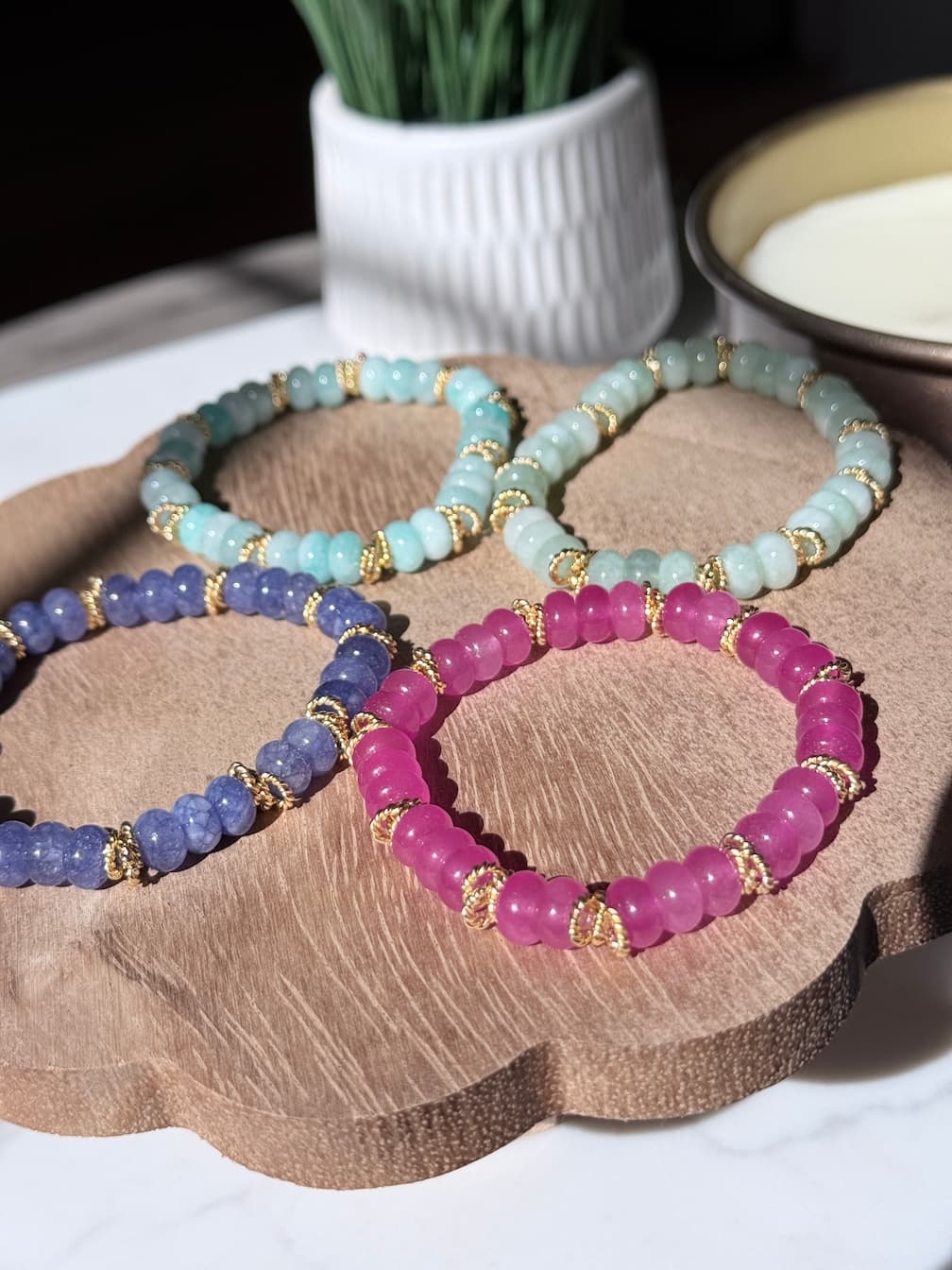 Four beaded stretch bracelets in light blue, lavender, and magenta, arranged on a scalloped wooden tray with gold twist spacer beads photographed in bright natural light with soft shadows.  A white ceramic pot and a bowl are softly blurred in the background.