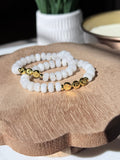 Two beaded stretch bracelets with white stone-like beads and gold alpha character beads, displayed on a wooden board with a blurred background.