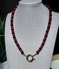 A beaded necklace with alternating glossy red and black beads, featuring a gold sailor clasp, displayed on a textured  jewelry bust.