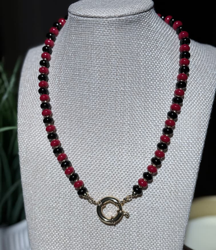 A beaded necklace with alternating glossy red and black beads, featuring a gold sailor clasp, displayed on a textured  jewelry bust.