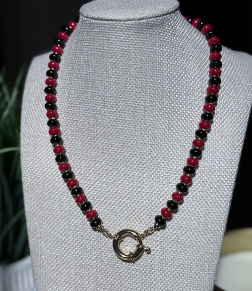A beaded necklace with alternating glossy red and black beads, featuring a gold sailor clasp, displayed on a textured  jewelry bust.