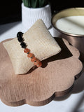 A beaded bracelet with black and orange beads displayed on a woven beige jewelry stand, set on a scalloped wooden tray with a candle and plant in the background.