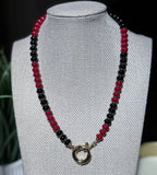 A beaded necklace with alternating red and black beads and a gold filled sailor clasp, displayed on a textured jewelry bust.