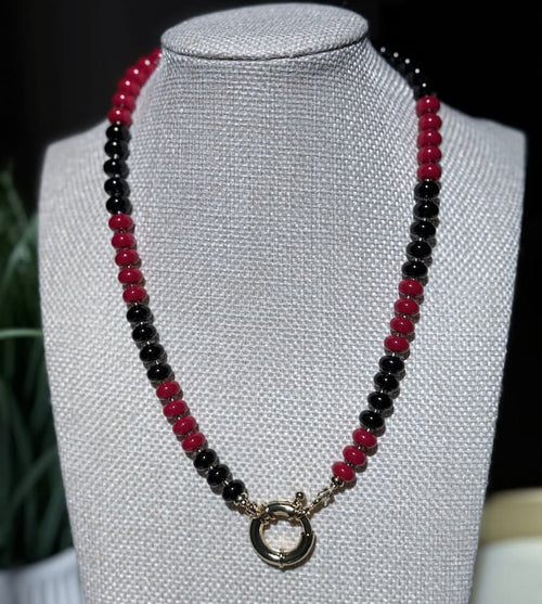 A beaded necklace with alternating red and black beads and a gold filled sailor clasp, displayed on a textured jewelry bust.