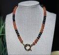 A beaded necklace with alternating strands of translucent orange and glossy black beads, featuring a gold-toned sailor clasp, displayed on a fabric jewelry bust.  To represent your favorite team colors.