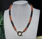 A beaded necklace with alternating strands of translucent orange and glossy black beads, featuring a gold-toned sailor clasp, displayed on a fabric jewelry bust.  To represent your favorite team colors.
