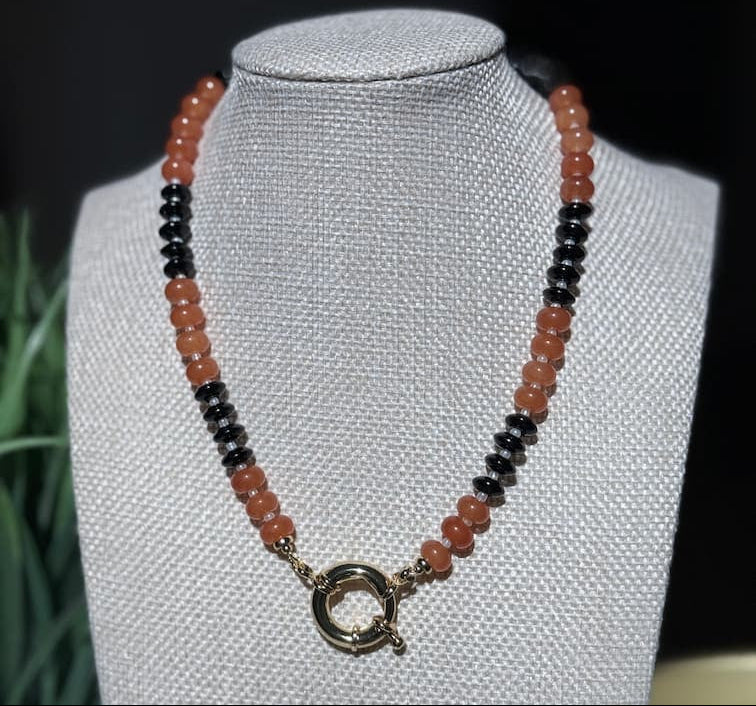 A beaded necklace with alternating strands of translucent orange and glossy black beads, featuring a gold-toned sailor clasp, displayed on a fabric jewelry bust.  To represent your favorite team colors.