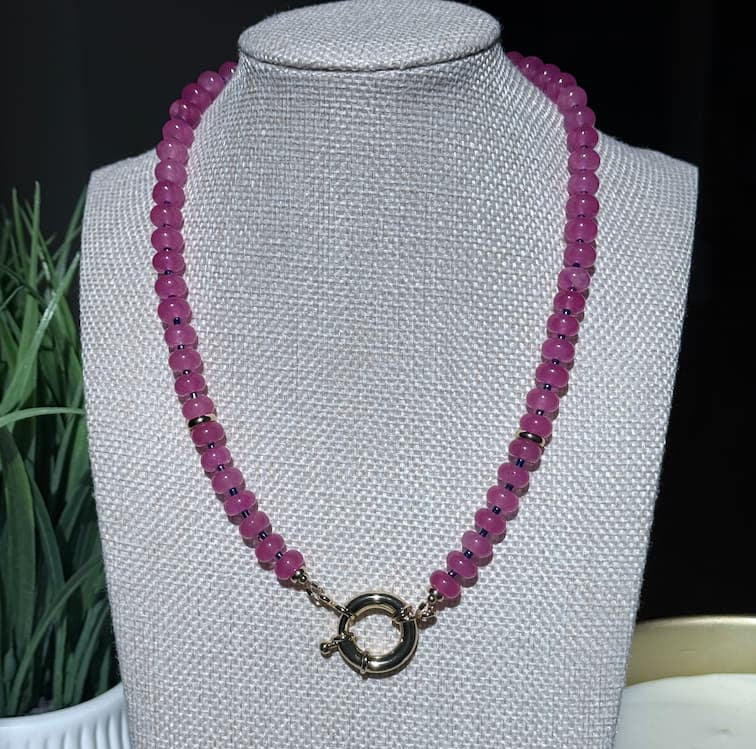 A handcrafted fuschia beaded necklace with a gold sailor clasp, displayed on a  jewelry bust.