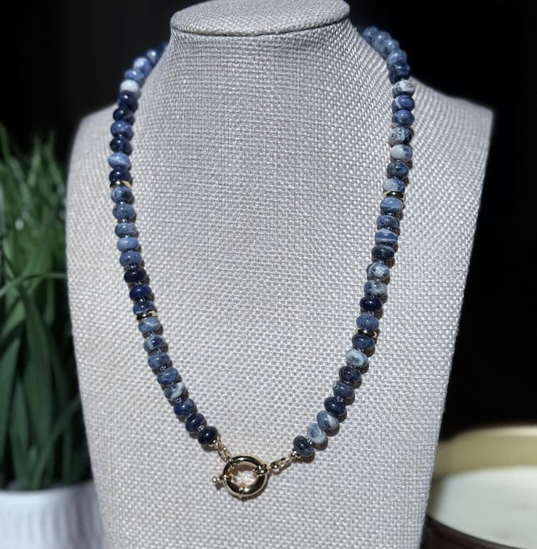 A beaded necklace with blue and gray stone beads and a small gold filled sailor clasp, displayed on a texturedjewelry bust against a dark background with blurred plant and candle props.