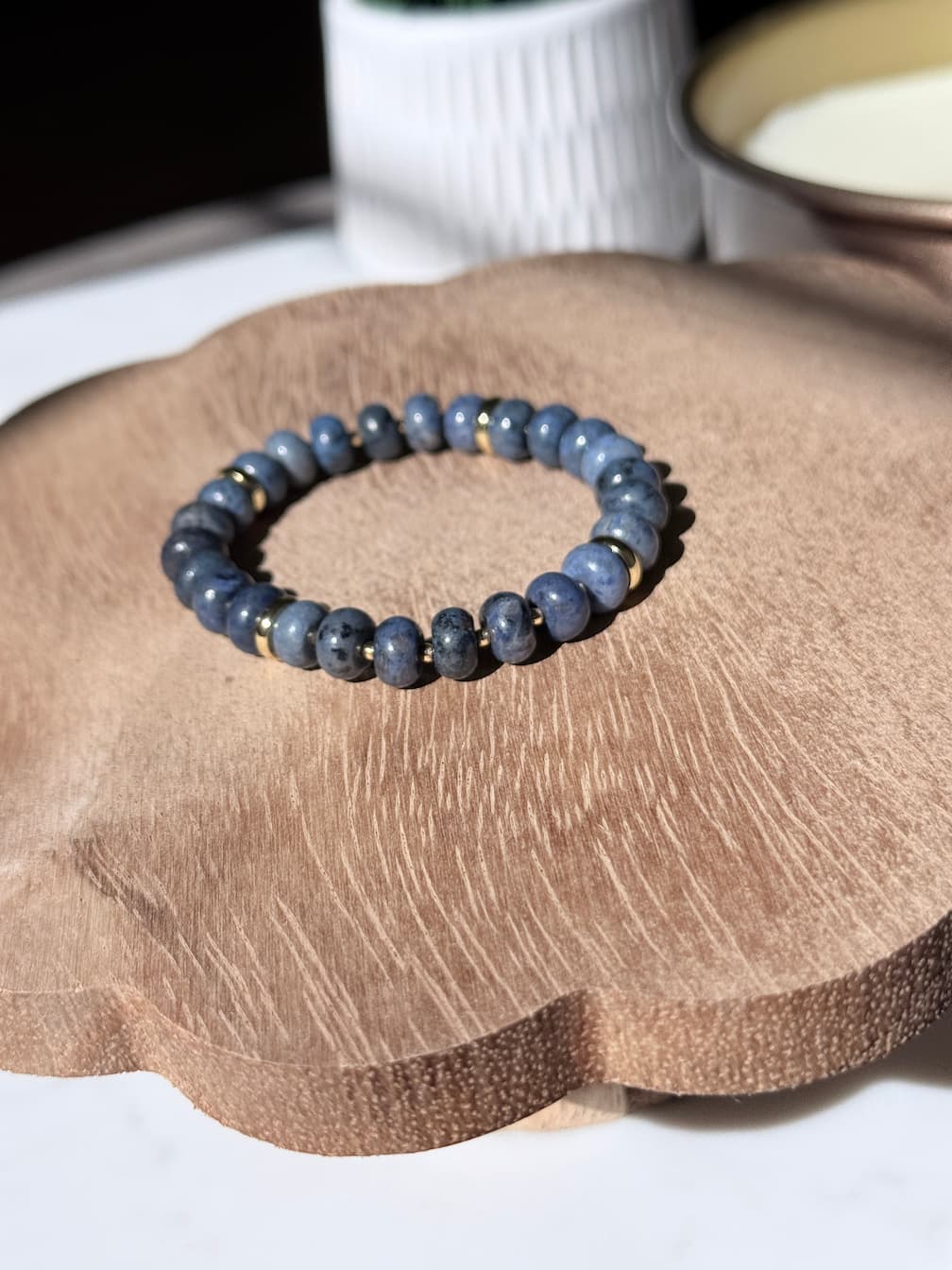 A beaded stretch bracelet made of dark blue-gray round beads with small gold spacer accents, displayed on a scalloped wooden tray.